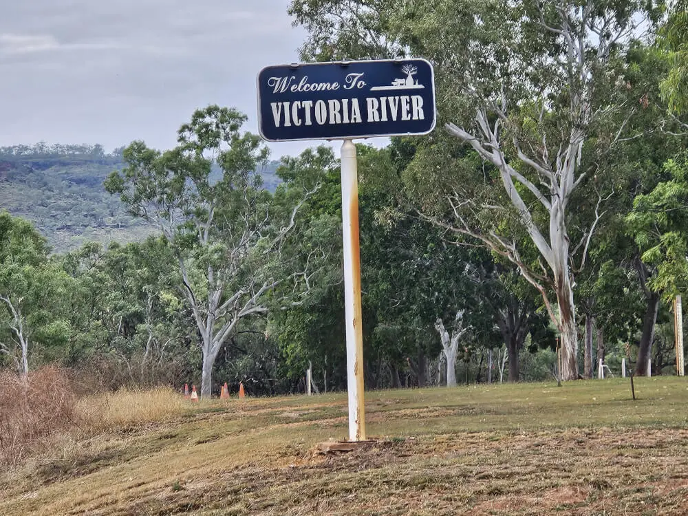 Welcome to Victoria River Blue Welcome to Victoria River sign beside a grassy roadside, with gum trees and rocky hills behind it.