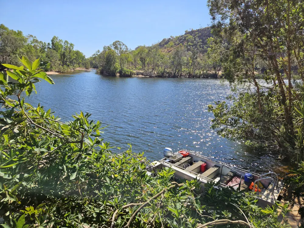 The Water at Katherine Gorge Calm blue water surrounded by trees and rocky hills with a small motorboat moored near the bank